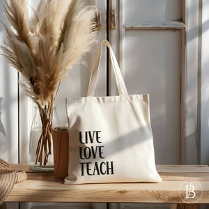 Könnte beinhalten: Eine weiße Canvas-Tragetasche mit schwarzem Text, der "Live Love Teach" lautet. Die Tasche liegt auf einer Holzoberfläche mit einer Vase mit getrocknetem Pampasgras im Hintergrund.