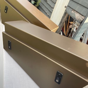 Puede incluir: Dos cajas metálicas rectangulares de color verde oliva con cierres negros. Las cajas están en ángulo, una apilada sobre la otra. Están al aire libre, sobre una superficie blanca, con una estructura de madera desgastada al fondo.