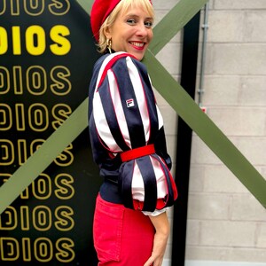 May include: A red beret worn with a navy blue and white long-sleeve top featuring a Fila logo and puffed sleeves with red accents, paired with red trousers. The top has a structured design with vertical navy stripes over white fabric.