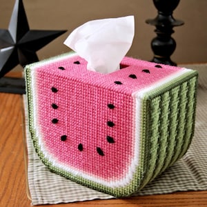 May include: A watermelon-shaped tissue box cover made of pink, green, and white yarn. The cover has black yarn details that resemble watermelon seeds.