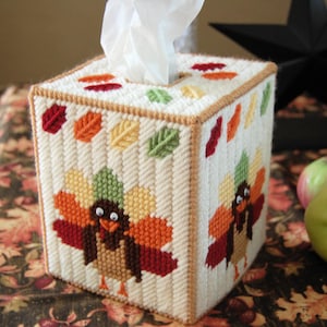 May include: A white tissue box cover with a Thanksgiving theme. The cover is decorated with embroidered turkeys and autumn leaves in shades of red, orange, yellow, and green.