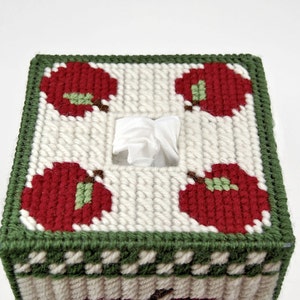 May include: A white tissue box cover with a green border and red apples with green leaves. The cover is made of yarn and has a square shape.