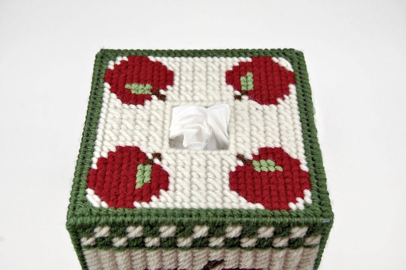 May include: A white tissue box cover with a green border and red apples with green leaves. The cover is made of yarn and has a square shape.
