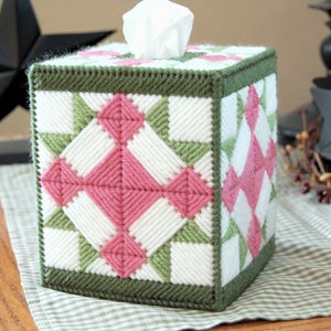 May include: A green, pink, and white tissue box cover with a quilted pattern. The cover is made of yarn and has a square shape.