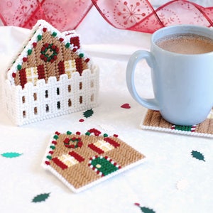 Puede incluir: Cuatro posavasos de crochet blanco con forma de casa de jengibre, uno apilado encima del otro, con una taza azul de chocolate caliente sobre un posavasos de casa de jengibre. Los posavasos están sobre una superficie blanca con corazones rojos y hojas verdes esparcidas alrededor.