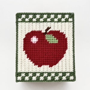 May include: A red apple with a green leaf and a white flower embroidered on a white canvas with a green and white checkered border.