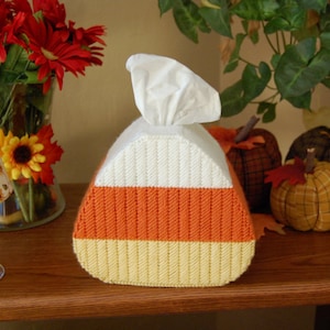 May include: A white, orange, and yellow candy corn-shaped tissue box cover. The cover is made of yarn and has a white tissue box inside.