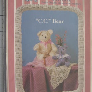 May include: A pink and white striped paper with the text "Cottage Crafts Patterns from Ozark Mountain Country" and a small illustration of a house. The paper also has a wicker frame with a blue background and the text "C.C. Bear" in black. The image shows two teddy bears, one white and one purple, sitting on a pink and white polka dot fabric with a white lace doily.