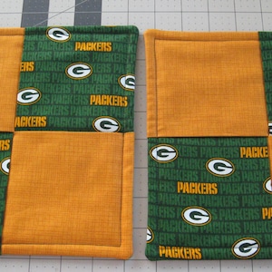 Op de afbeelding: Twee groene en gouden stoffen ovenwanten met een geruit patroon. De stof heeft de woorden "Packers" en het logo van de Green Bay Packers erop herhaald.