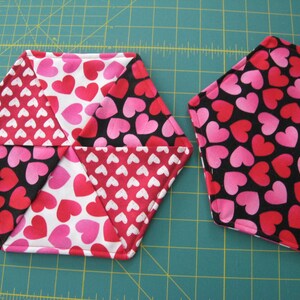May include: Two hexagonal fabric coasters with a heart pattern. One coaster is black with pink and red hearts, the other is white with pink and red hearts.