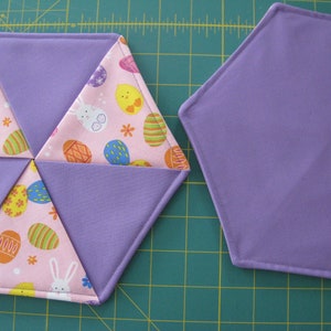 Könnte beinhalten: Zwei lila Stoff-Sechsecke, einer mit einem pinkfarbenen und weißen Stoffzentrum mit einem Muster aus Küken, Hasen und Ostereiern. Der andere Sechseck ist einfarbig lila.