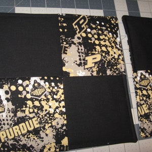 May include: A black and gold fabric patchwork with the word "Purdue" repeated in a stylized font. The fabric has a splatter paint design with gold circles and black and white accents.