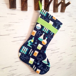 May include: A Christmas stocking with a navy blue background and a science-themed pattern. The pattern includes beakers, test tubes, and molecular structures in shades of teal, orange, and white. A bright green band and hanging loop accent the top.