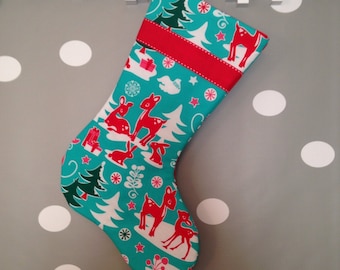 May include: A Christmas stocking with a teal background and a red cuff. The stocking is decorated with red deer, white trees, and gift boxes. A red ribbon is attached for hanging.