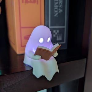 May include: A small, white, ghost-shaped LED lamp with a soft purple glow, featuring two bright white circular eyes and holding a small, brown, open book in its hands. The figure is positioned to sit on the edge of a dark shelf.