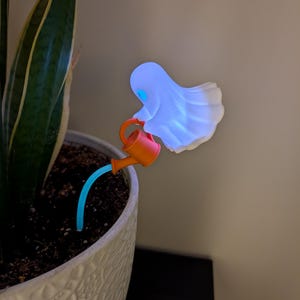 May include: A decorative plant stake featuring a glowing white ghost figure holding a small orange watering can. A thin, light blue stem extends from the watering can into the soil, creating a whimsical scene of a ghost watering a plant.