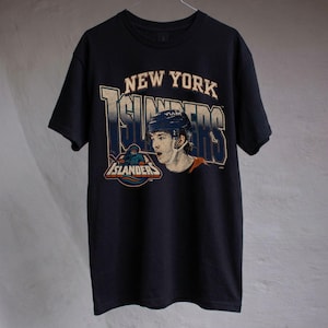 May include: A black short-sleeved t-shirt featuring a graphic print of a hockey player wearing a blue helmet. The text reads NEW YORK ISLANDERS in large block letters above the player, with an additional ISLANDERS logo positioned to the left.