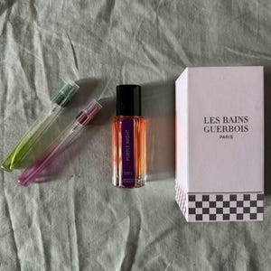 Puede incluir: Un frasco de perfume con tapa negra y etiqueta morada que dice "Purple Night 1992" junto a una caja blanca con "Les Bains Guerbois Paris". También hay dos frascos de spray de vidrio transparente con tapas verdes y moradas.