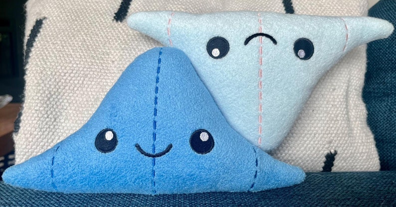 May include: Two plush toys shaped like manta rays. The blue manta ray is smiling with black eyes. The light blue manta ray has a sad expression with black eyes.