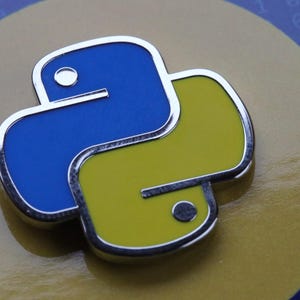 以下が含まれることがあります： 青と黄色の Python ロゴがついた銀色の金属製のピン。ロゴは、青い頭と黄色の体を持つスタイリッシュなヘビです。