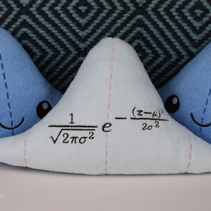 May include: Three blue plush triangle pillows with embroidered faces. The middle pillow has a mathematical formula embroidered on it: "1/&radic;2&pi;&sigma;&sup2; e^-(x-&mu;)&sup2;/2&sigma;&sup2;".