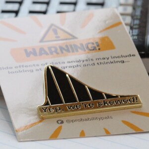 May include: A gold-colored pin with a black and gold skewed graph design. The pin has the text "Yep, we're skewed!" and "@probabilitypals" on it. The pin is on a white card with a yellow warning sign and the text "WARNING! Side effects of data analysis may include looking at graph and thinking..."