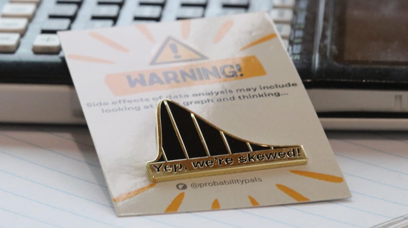 May include: A gold-colored pin with a black and gold skewed graph design. The pin has the text "Yep, we're skewed!" and "@probabilitypals" on it. The pin is on a white card with a yellow warning sign and the text "WARNING! Side effects of data analysis may include looking at graph and thinking..."