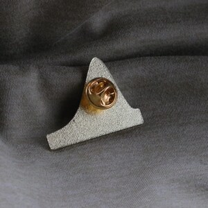 May include: The back of a silver metal pin with a gold-colored pin back. The pin is shaped like a triangle with a rounded top.