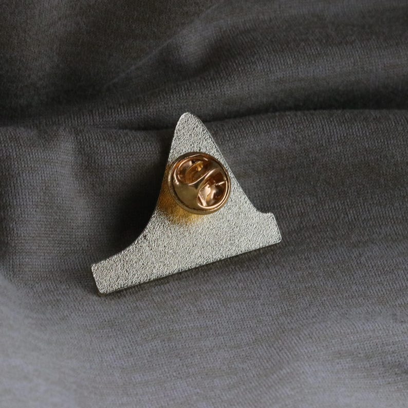 May include: The back of a silver metal pin with a gold-colored pin back. The pin is shaped like a triangle with a rounded top.