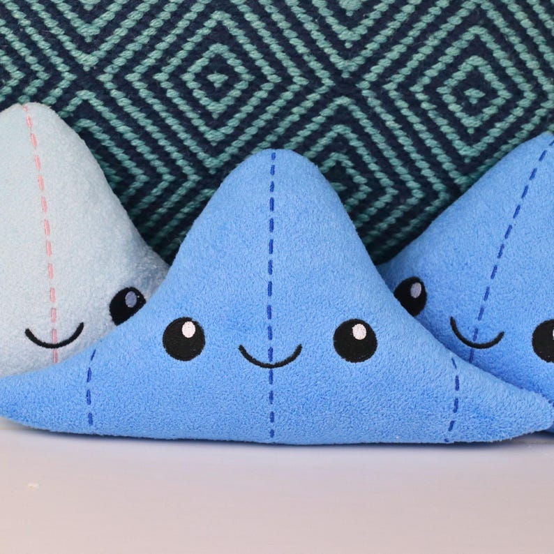 May include: Three plush blue and light blue star-shaped pillows with embroidered eyes and smiling mouths. The pillows are arranged in a row on a white surface.