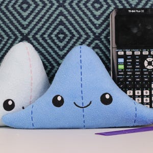 May include: Two blue and light blue plush toys shaped like triangles with embroidered eyes and a smiling mouth. The toys are sitting on a blue and white patterned surface next to a black calculator with the text "TI-84 Plus CE".
