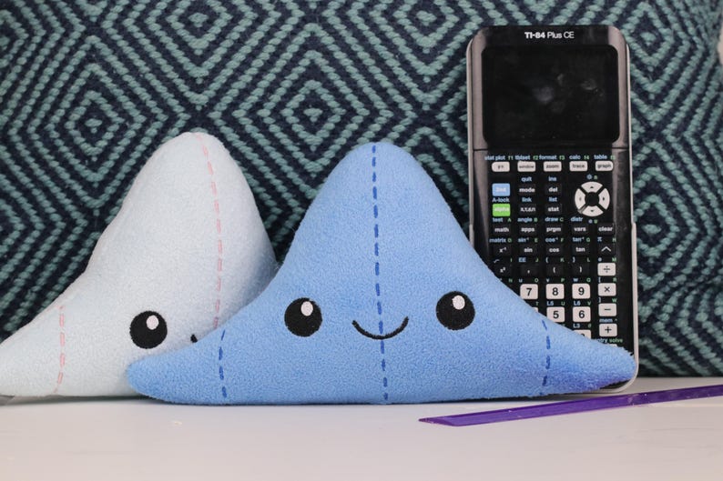 May include: Two blue and light blue plush toys shaped like triangles with embroidered eyes and a smiling mouth. The toys are sitting on a blue and white patterned surface next to a black calculator with the text "TI-84 Plus CE".
