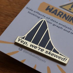 Puede incluir: Pin de esmalte dorado con una curva de campana negra sesgada y el texto "Yep, we're skewed!" y "@probabilitypals". El pin está en una tarjeta con el texto "Side effects of data analysis may include looking at our graph and thinking... WARNING!"