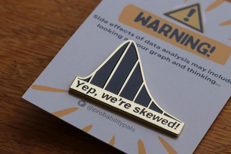 May include: Gold-colored enamel pin with a black skewed bell curve and the text "Yep, we're skewed!" and "@probabilitypals". The pin is on a card with the text "Side effects of data analysis may include looking at our graph and thinking... WARNING!"