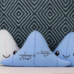 May include: Three blue and white plush pillows shaped like triangles. The middle pillow has a mathematical formula embroidered on it: "1/&radic;2&pi;&sigma;&sup2; e^-(x-&mu;)&sup2;/2&sigma;&sup2;".