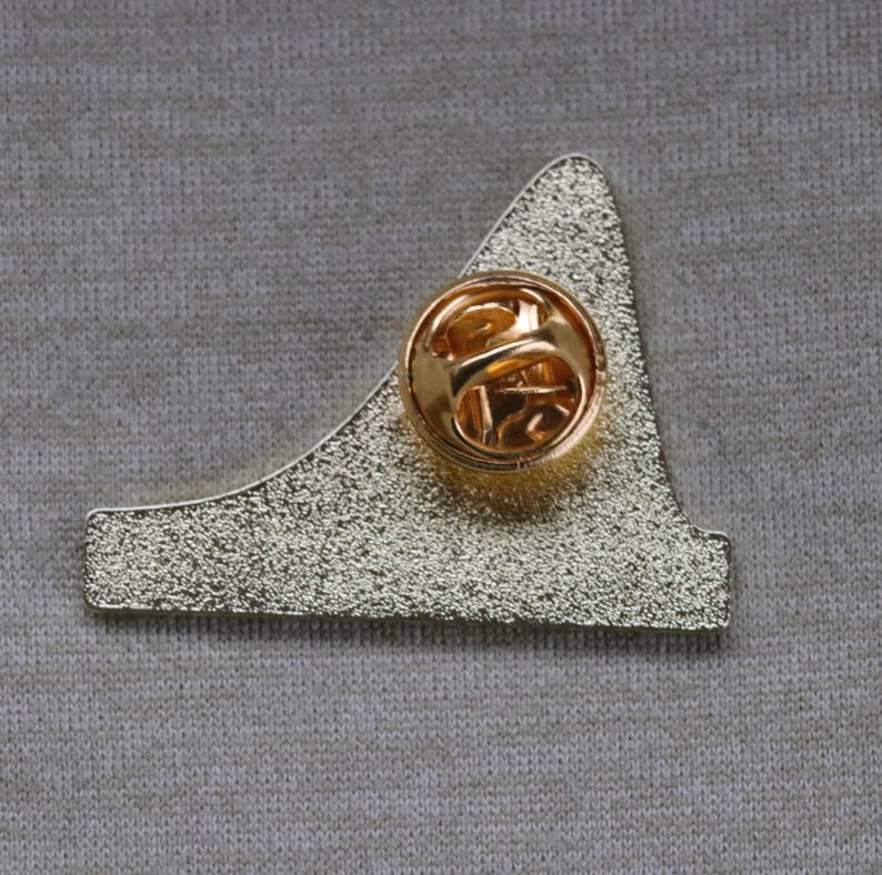 May include: Gold-colored metal pin with a geometric shape. The back of the pin has a standard pin back.
