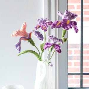 May include: A white vase holds a bouquet of handmade crochet flowers in shades of purple, pink, and green. The vase sits on a wooden table next to a candle and a window with a brick exterior visible.
