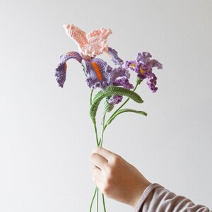 May include: A bouquet of handmade crochet flowers, featuring purple, pink, and orange irises with green stems and leaves. The flowers are held in a person's hand against a plain white background.