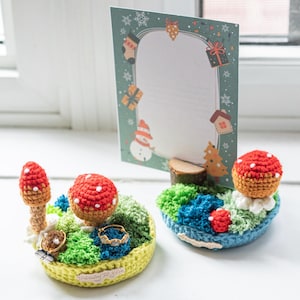 May include: Two handmade crochet ring holders shaped like miniature mushroom gardens. One is yellow, green, and blue, and the other is blue, green, and white. A Christmas card with a snowman and gifts is in the background.