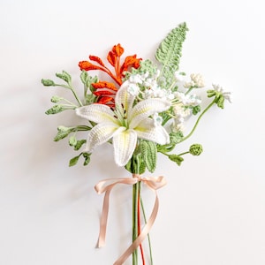 May include: A handmade crochet bouquet featuring a large white lily with yellow detailing, bright orange flowers, and small white blooms. The bouquet includes green crocheted leaves and stems, tied with a soft pink ribbon. A unique, handcrafted floral arrangement.