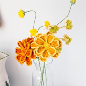 May include: A clear glass vase holds a bouquet of handmade crochet flowers. The flowers are in shades of yellow and orange, with green stems. The crochet flowers have detailed petal designs, creating a textured appearance. The vase sits on a gold wire rack.