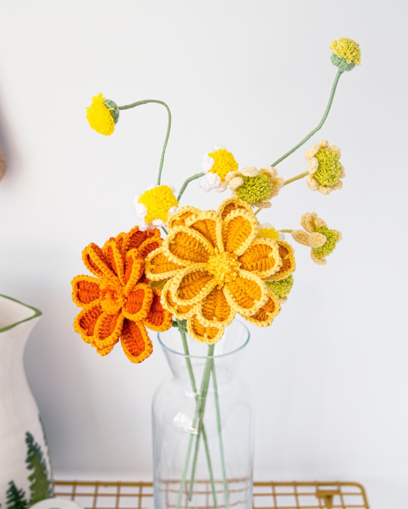 May include: A clear glass vase holds a bouquet of handmade crochet flowers. The flowers are in shades of yellow and orange, with green stems. The crochet flowers have detailed petal designs, creating a textured appearance. The vase sits on a gold wire rack.