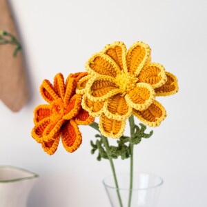 May include: Two handmade crochet flowers in a clear glass vase. One flower is bright orange, the other is yellow with orange edging. Green stems and leaves support the flowers. The flowers are set against a white background.