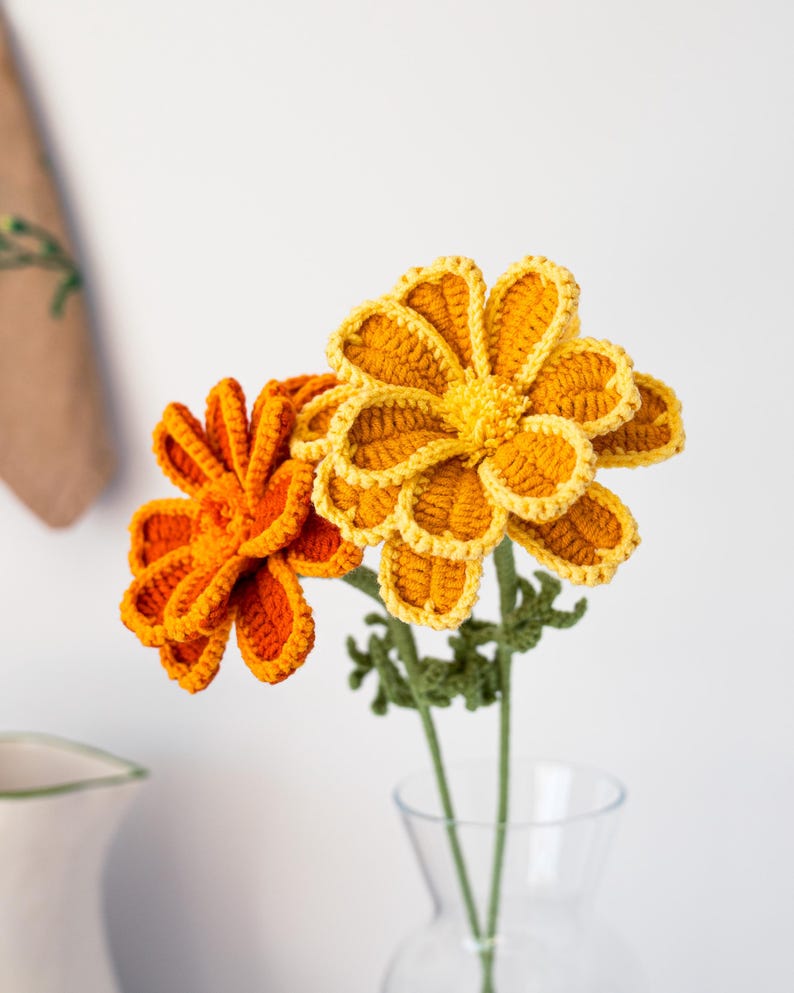 May include: Two handmade crochet flowers in a clear glass vase. One flower is bright orange, the other is yellow with orange edging. Green stems and leaves support the flowers. The flowers are set against a white background.