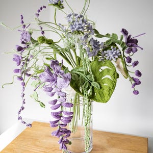 May include: A crocheted bouquet of purple and green flowers in a clear glass vase. The flowers are arranged in a cascading style, with the purple flowers hanging down from the vase.