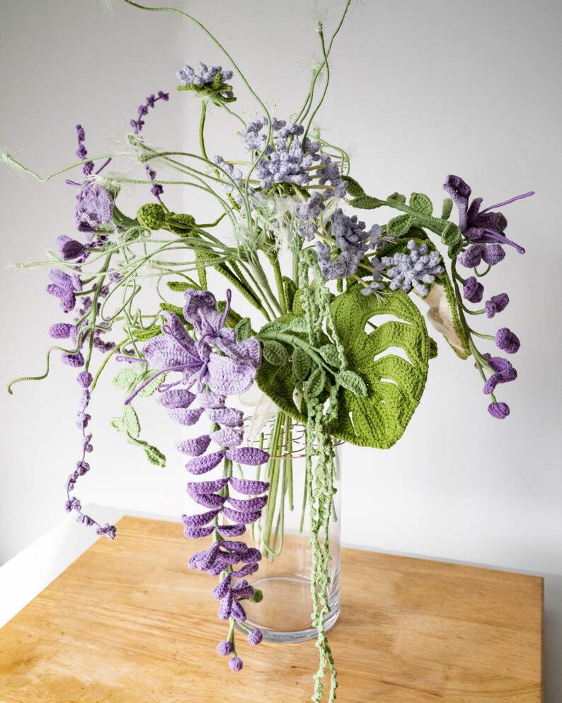 May include: A crocheted bouquet of purple and green flowers in a clear glass vase. The flowers are arranged in a cascading style, with the purple flowers hanging down from the vase.