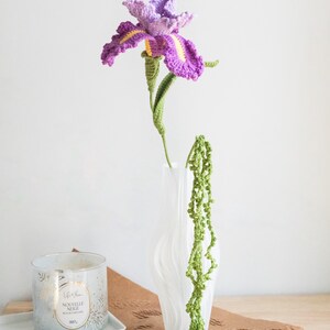 May include: A handmade crochet iris flower in shades of purple and yellow, with a green stem, displayed in a white vase. A green crochet vine hangs from the vase. A candle in a glass jar sits on a white tray.