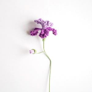 May include: A handmade crochet flower in shades of purple and lavender, with a green stem and a bud. The flower has detailed petals and a textured appearance, set against a white background. This decorative item is a unique craft.