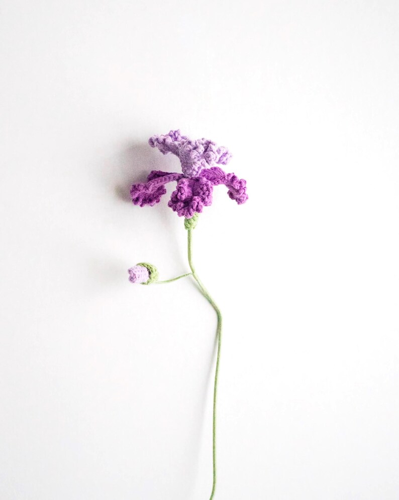 May include: A handmade crochet flower in shades of purple and lavender, with a green stem and a bud. The flower has detailed petals and a textured appearance, set against a white background. This decorative item is a unique craft.
