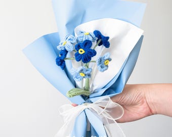 May include: A small bouquet of blue and yellow crocheted forget-me-not flowers wrapped in light blue tissue paper with a white ribbon. The bouquet is tied with a white ribbon and has a green stem. The text "the way to you" is written on the paper.